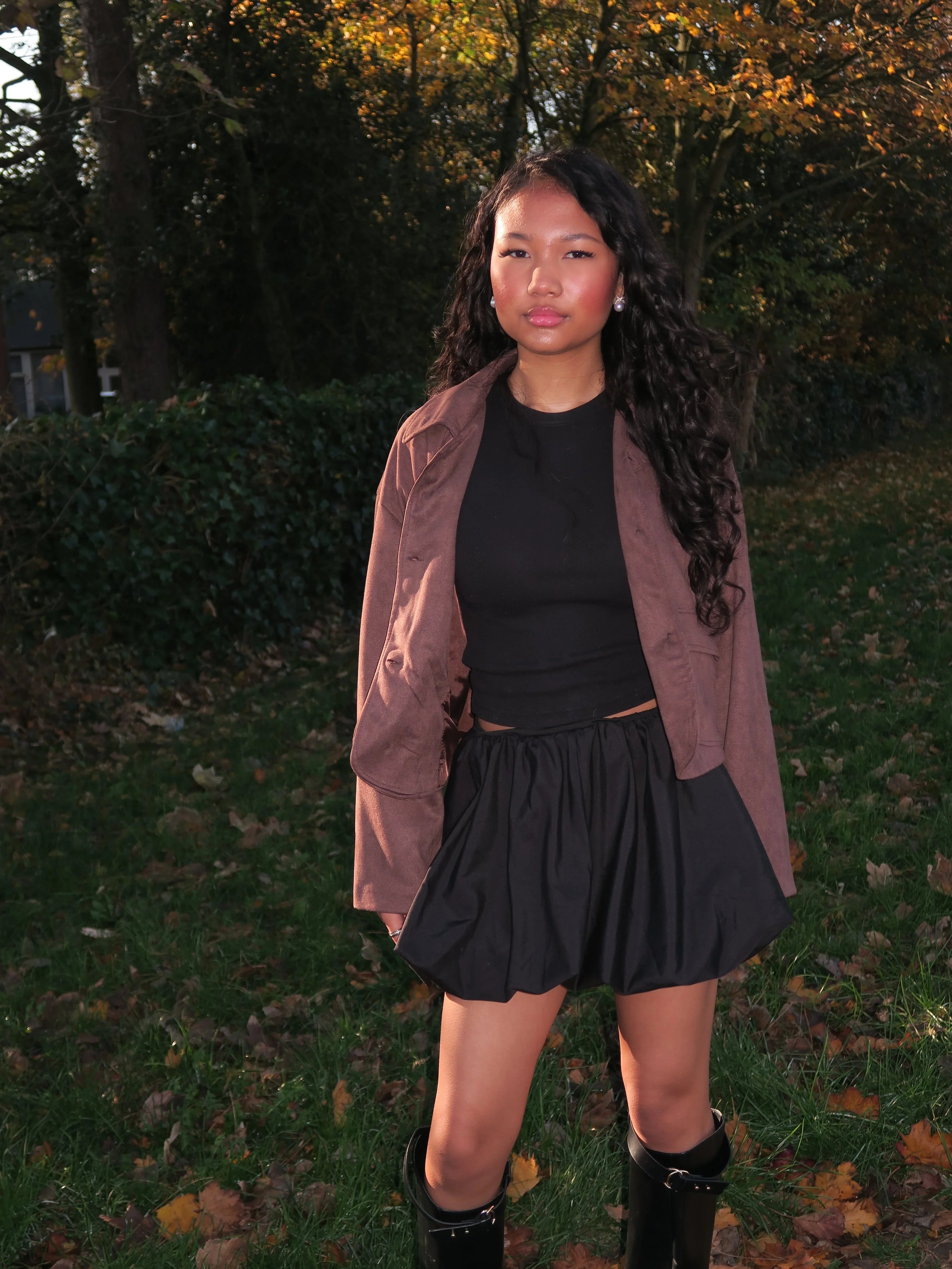 Young woman in black crop top, black skirt, brown jacket, and black boots standing outdoors in autumn