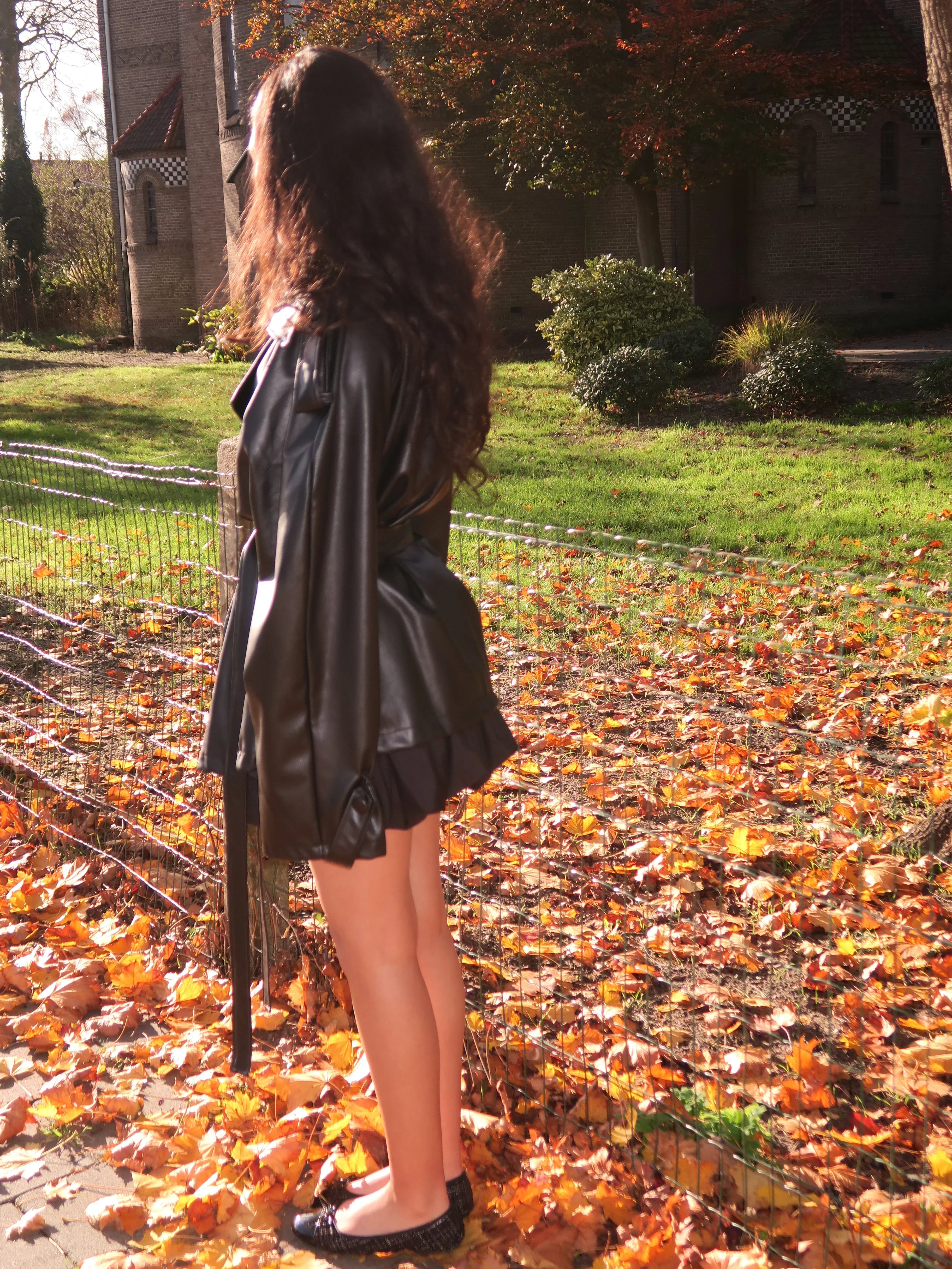 Woman wearing black leather jacket and flats standing on autumn leaves near a park fence