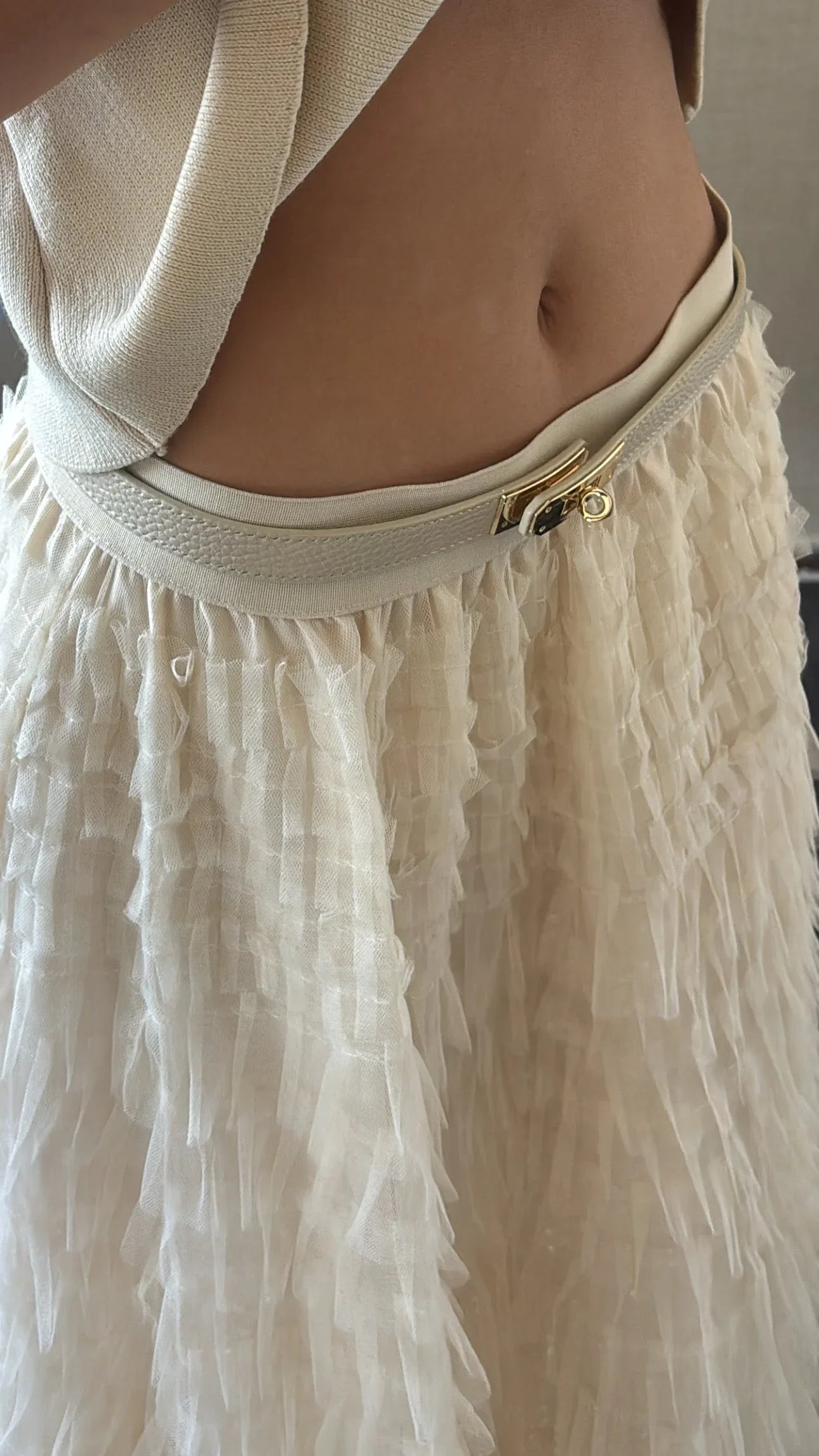 Close-up of woman's waist wearing a beige knit top with a cream tiered ruffle skirt and gold buckle belt