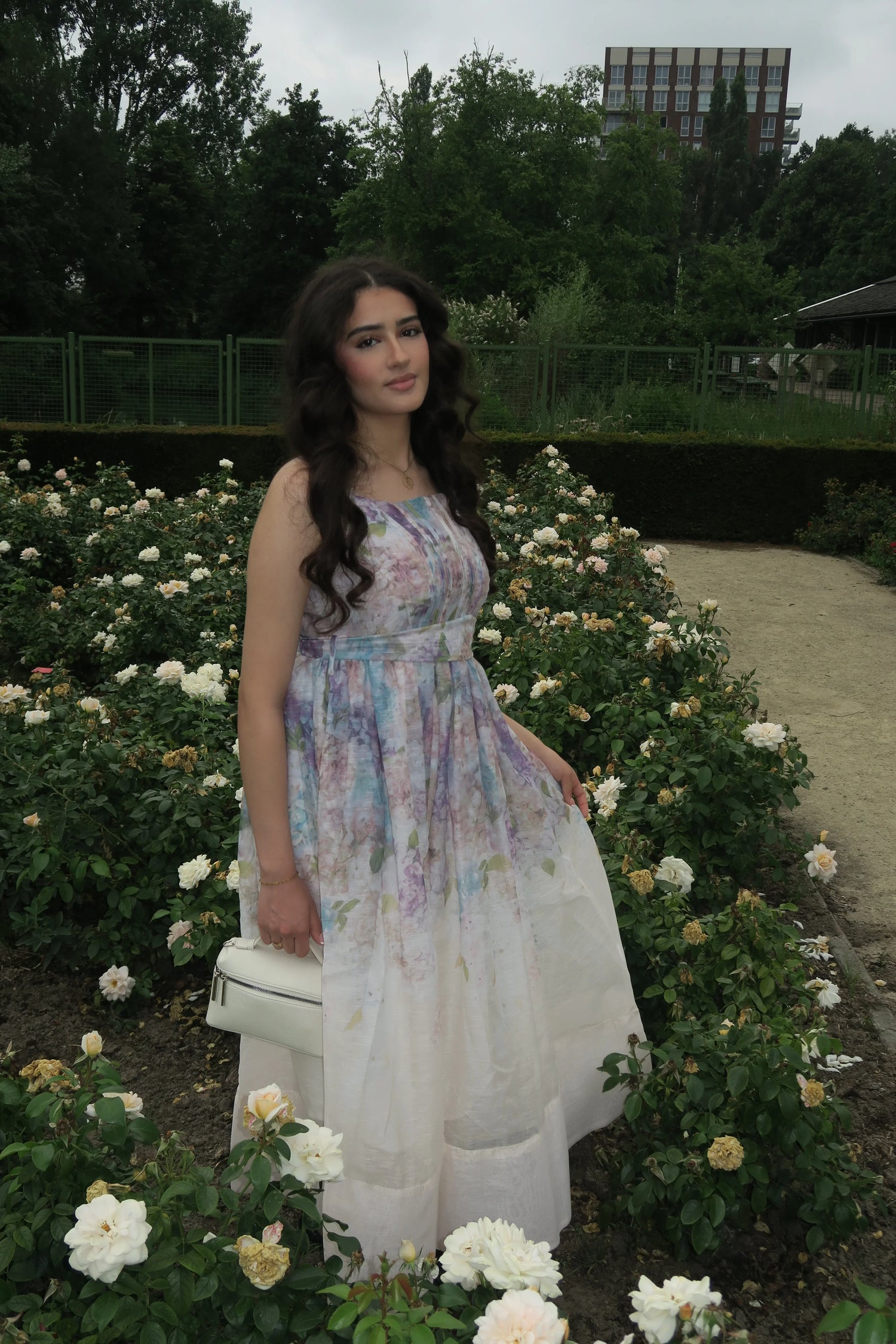 Woman in floral pastel maxi dress holding white handbag, standing in rose garden with greenery background
