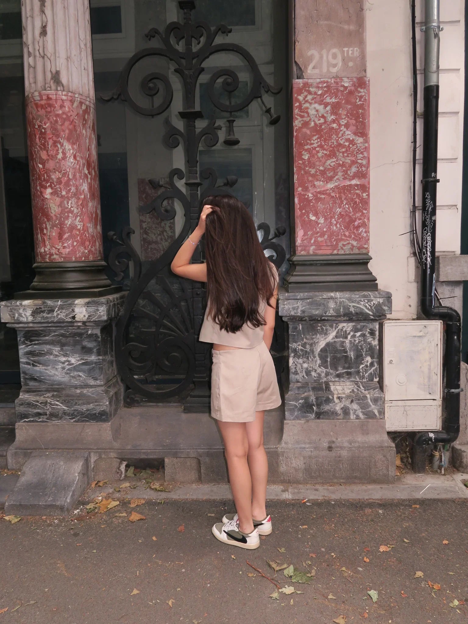Woman with long brown hair in beige outfit standing by decorative iron gate and marble pillars
